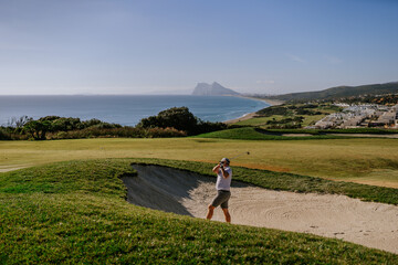 Fototapeta na wymiar Sotogrande, Spain - January, 23, 2024 - Golfer playing a shot from a sand bunker with the Rock of Gibraltar and Mediterranean coastline in the background.