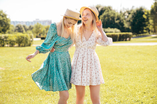 Two young beautiful smiling hipster woman in trendy summer dress. Carefree women in the street in hats. Positive models at sunset. Cheerful and happy. Walking barefoot, enjoy summertime days