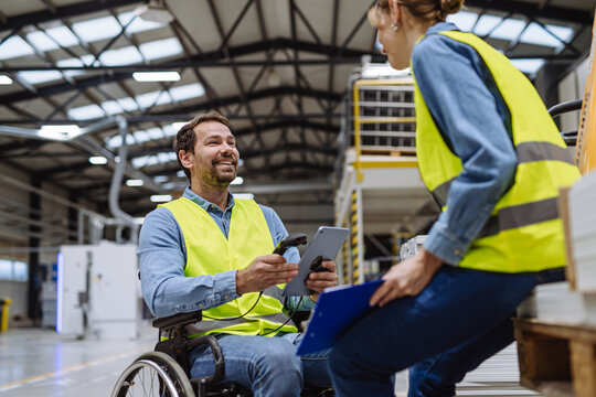 Portrait of man in wheelchair working in modern industrial factory, talking with female coworker. Concept of workers with disabilities, accessible workplace for employees with mobility impairment. - Powered by Adobe