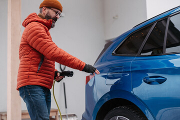 Man charging electric car during cold snowy day. Side view of hansome man putting charger in charging port during winter, cold weather. Charging and driving electric vehicles during winter season.
