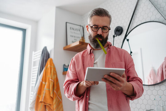 Man shopping online while brushing his teeth in bathroom. Holding tablet, scrolling and buying in eshop, toothbrush in mouth.