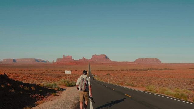 Camera follows traveler man with backpack and camera walking along hot sunny straight desert road at Arizona slow motion