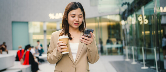 Young asian elegant business woman leader wearing suit standing in modern city building using cell phone platform applications hold coffee drink