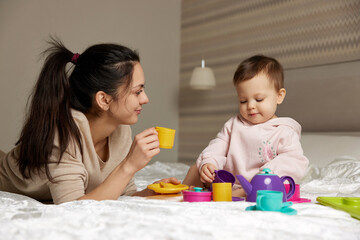 happy mother and little child daughter pretending drinking tea from plastic toy cups and spending time together in bedroom
