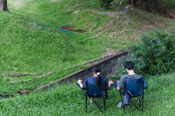 young couple sat happily chatting on camping chairs he had prepared on bright green lawn and beautiful nature background. A young couple happily sits and chats amidst beautiful nature.