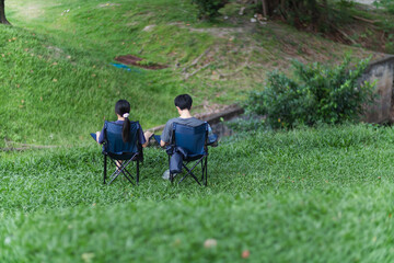 young couple sat happily chatting on camping chairs he had prepared on bright green lawn and beautiful nature background. A young couple happily sits and chats amidst beautiful nature.