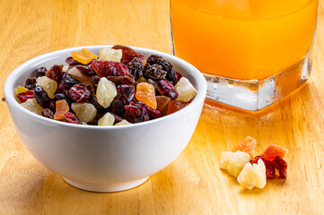 Closeup of various dried tropical fruit in white ceramic bowl and orange juice in transparent glass on wooden board.