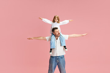 Father and daughter with arms outstretched, play airplanes pretending to fly against pink pastel...