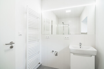 A Bright, clean bathroom with a white sink, chrome faucet, large mirror, and a visible shower enclosure. A white door is on the left.