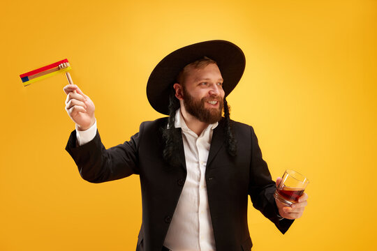 Cheerful young Jewish man in hat, with sidelocks holding noisemaker, against yellow background. Celebration. Concept of Purim holiday, Jewish traditions, history and culture