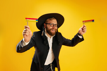 Bearded young Jewish man in glasses, yarmulke playing with wooden noisemaker, gragger against...
