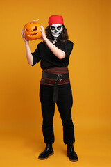 Man in scary pirate costume with skull makeup and carved pumpkin on orange background. Halloween celebration
