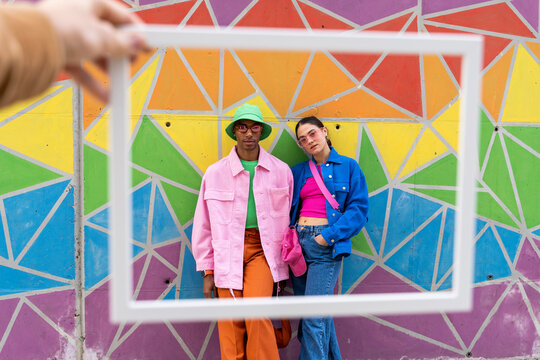 Cropped hand holding picture frame with friends standing in front of colorful wall