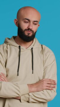 Vertical Video Young adult in a hoodie waiting for something in studio, standing with arms crossed over blue background. Middle eastern man being impatient on camera, punctual tensed person. Camera 1.
