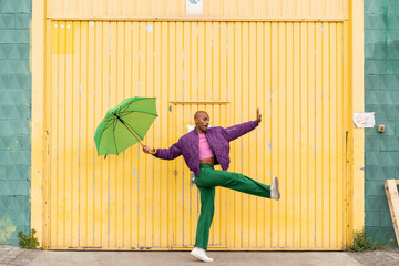 Non-binary person dancing with green umbrella in front of yellow shutter door