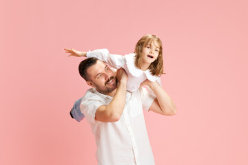 Man with playful smile carrying young girl on his shoulders, both simulating flight against pink pastel background. Concept of International Day of Happiness, childhood and parenthood. Ad - obrazy, fototapety, plakaty