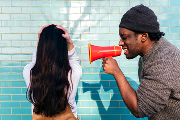 Angry man shouting through megaphone on woman with head in hands facing wall