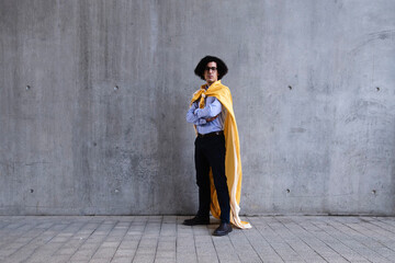 Man wearing yellow cape standing with arms crossed in front of gray wall