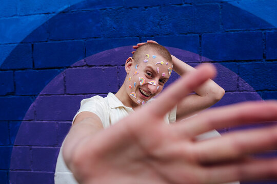 Happy non binary person with stickers on face showing stop gesture in front of colorful wall