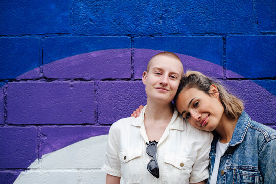 Happy woman leaning head on non binary friend's head in front of wall