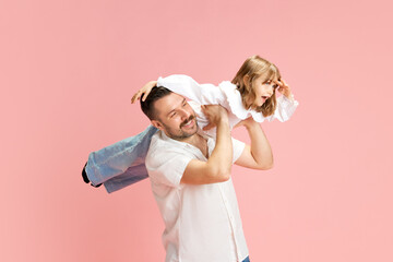 Cheerful father playing with daughter holding her on shoulder acting as airplane against pink pastel background. Concept of International Day of Happiness, childhood and parenthood, positive emotions