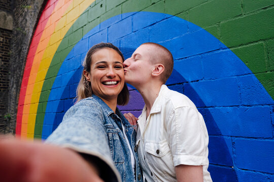 Non binary person kissing friend in front of rainbow wall