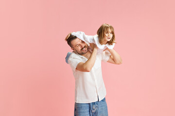 Joyful dad and child engaged in imaginative play, simulating flying against pink pastel background. Concept of International Day of Happiness, childhood and parenthood, positive emotions. Ad