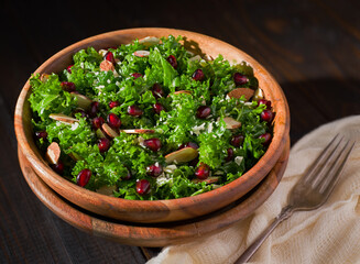 Healthy organic kale salad with pomegranate, slivered almonds, and Parmesan cheese in a wooden bowl on a dark wooden surface; copy space