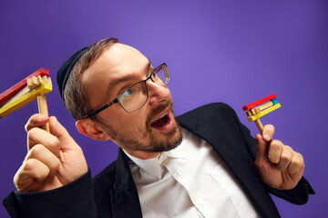 Cheerful Jewish man in yarmulke and glasses holding noisemaker, against purple studio background....