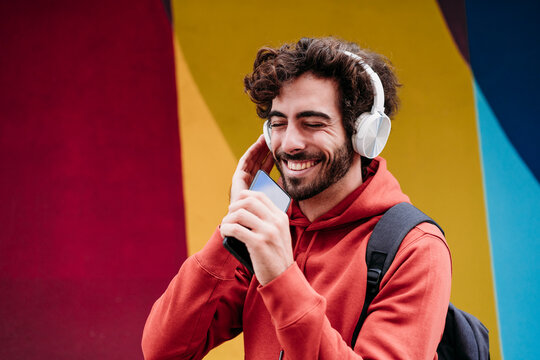 Happy man with eyes closed listening to music holding smart phone in front of colorful wall