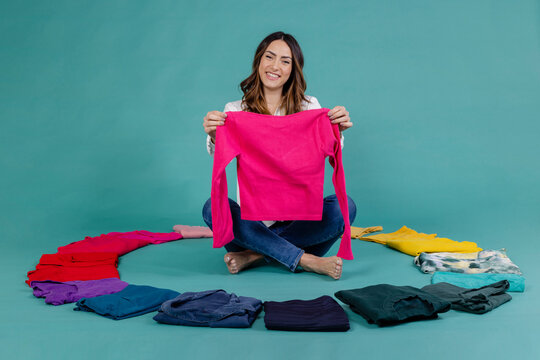 Smiling woman holding pink t-shirt sitting cross-legged amidst clothes against blue background
