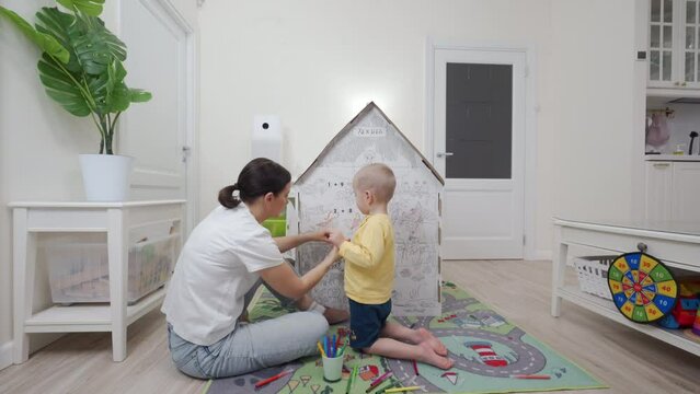 mother with child playing with house cardboard coloring playhouse, little boy drawing on wall of carton house toy for children, kid having fun at home.