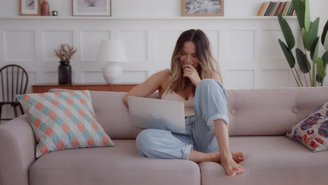 young brunette woman sits in couch in living room during daylight, smiles and laughs looking at screen, feels delighted and pleased on home weekend