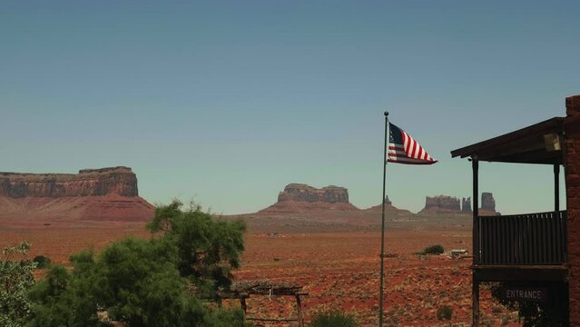 Dusty American flag waving in the middle of Monument Valley sunny hot desert scenery and old wooden Wild West building.
