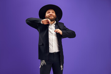 Emotional young Jewish man in hat, with sidelocks holding dancing against purple studio background....