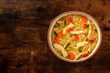 Chicken noodle soup with vegetables, a bowl of healthy broth, shot from above on a rustic wooden table, winter comfort food with copy space