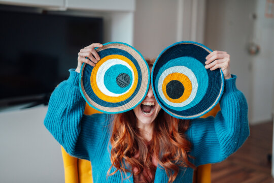 Happy woman holding macrame evil eyes over face at home