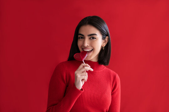 Happy young woman eating heart shaped lollipop against red background