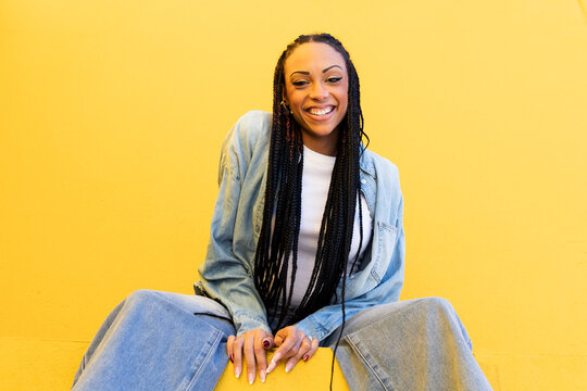 Happy woman with box braids sitting near yellow wall