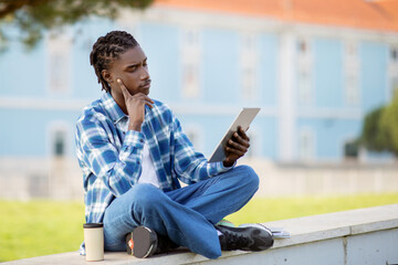 Thoughtful African student guy engages in e-learning on tablet outside