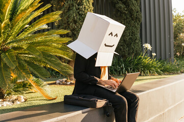 Freelancer wearing box with smiley face using laptop sitting on wall