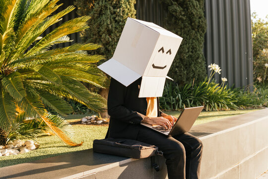 Freelancer wearing box with smiley face using laptop sitting on wall