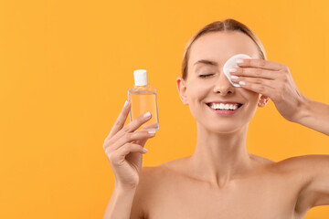 Smiling woman removing makeup with cotton pad and holding bottle on yellow background. Space for text