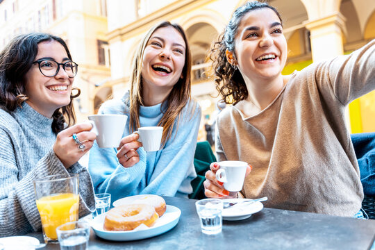 Happy smiling young women enjoying breakfast drinking coffee at bar cafeteria - Group of female friends taking selfie picture with smart mobile phone outside