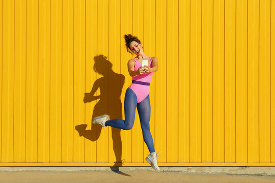 Happy woman dancing and taking selfie through smart phone in front of yellow wall