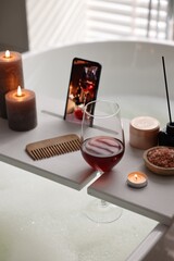 White wooden tray with smartphone, glass of wine and burning candles on bathtub in bathroom