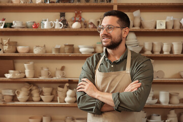 Portrait of smiling craftsman near shelf with different pottery in workshop. Space for text
