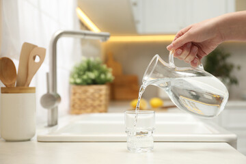 Woman pouring water from jug into glass at white table in kitchen, closeup. Space for text