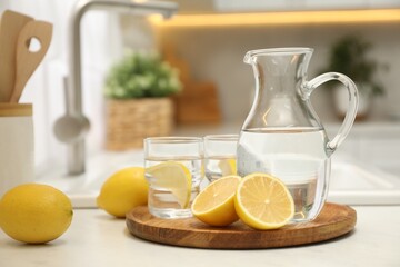 Jug, glasses with clear water and lemons on white table in kitchen