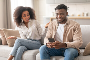 Discontented Black Wife Looking At Husband Browsing On Phone Indoor